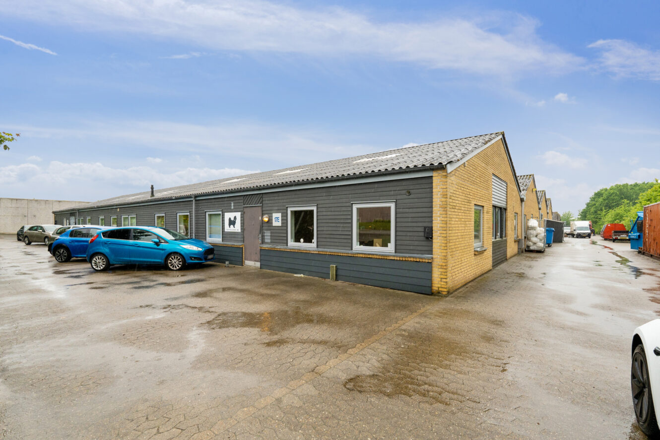 Industribygning i én etage med gule mursten og grå facadebeklædning, flere parkerede biler og vådt fortov under en delvis overskyet himmel.