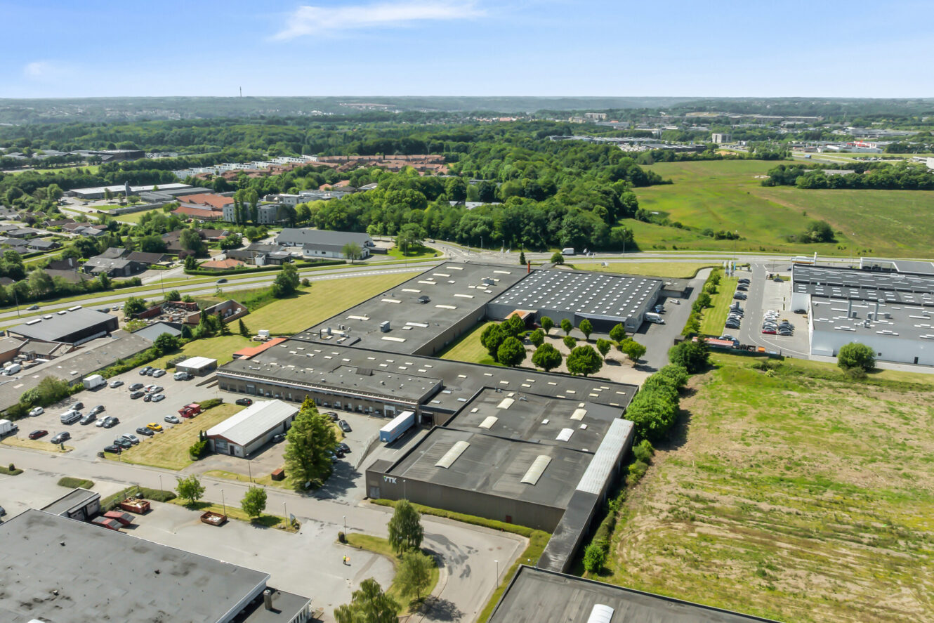 Luftfoto af et industrikompleks med flere store lagerbygninger, parkeringspladser og omkringliggende grønne marker under en klar himmel.