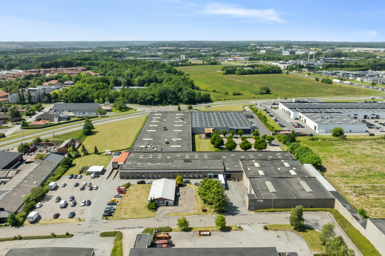 Luftfoto af et industrikompleks med flere store lagerbygninger, parkeringspladser og omkringliggende grønne områder i et forstadsområde.