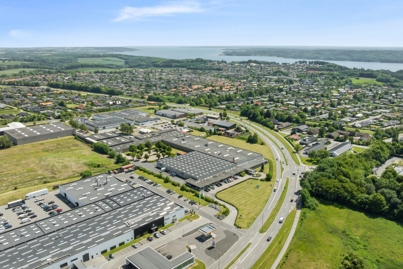 Luftfoto af et industriområde med lagerbygninger, veje, parkerede biler og tilstødende boligkvarterer med en vandmasse og grønne områder i baggrunden.