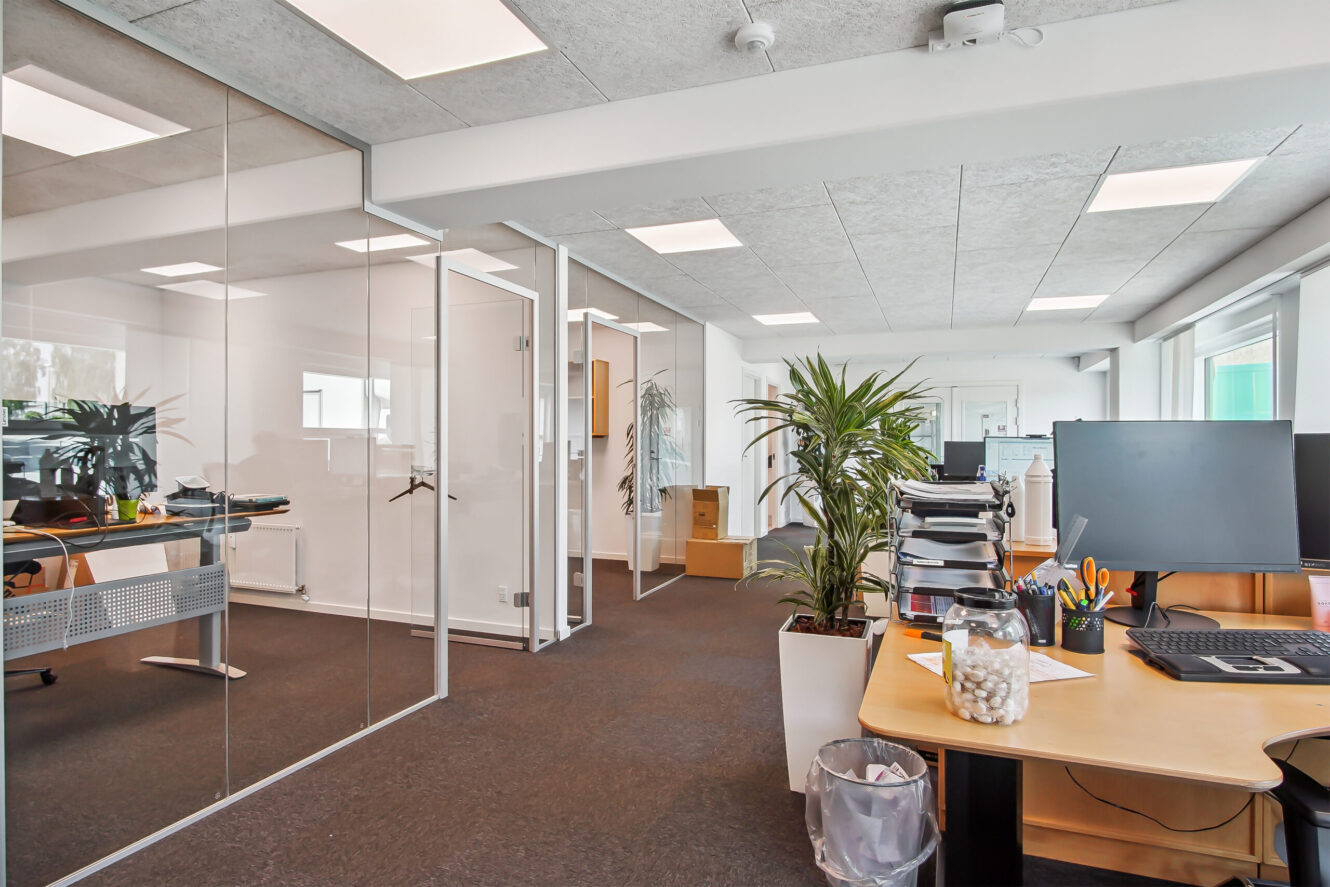 Moderne kontorlokaler med skriveborde, computere, glasvægge, kontorartikler, potteplanter og naturligt lys fra store vinduer.