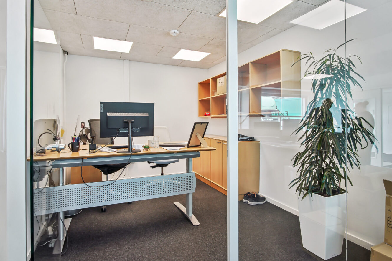 Moderne kontor med skrivebord, computerskærm, stol, hylder, en potteplante og glasvægge, set udefra.
