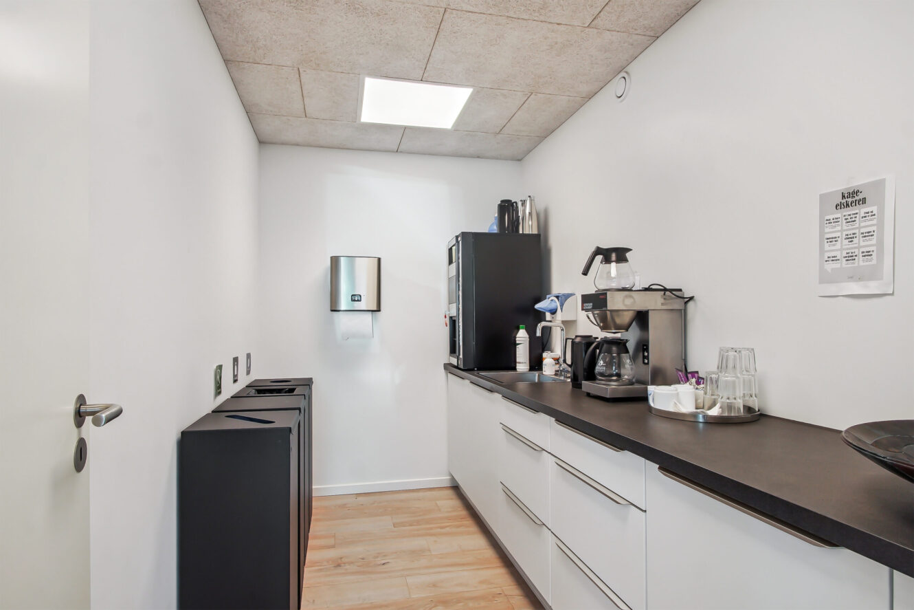 Et lille, moderne tekøkken med hvide skabe, sorte bordplader, et køleskab, kaffemaskiner, en papirhåndklædedispenser og en affaldsspand mod hvide vægge.