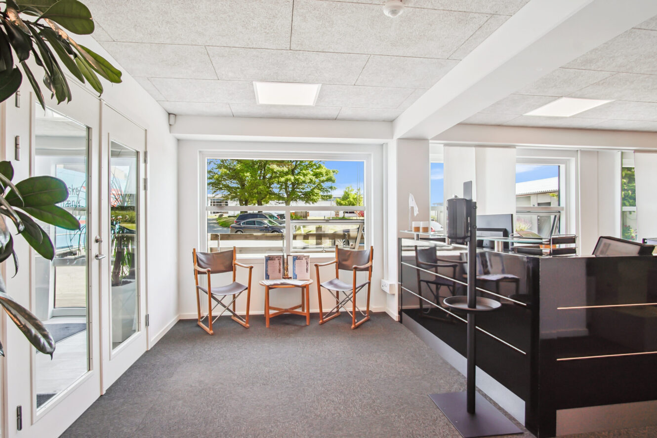 Moderne kontorindretning med to stole og et lille bord ved store vinduer, sort receptionsskranke til højre og planter ved glasdøren til venstre.