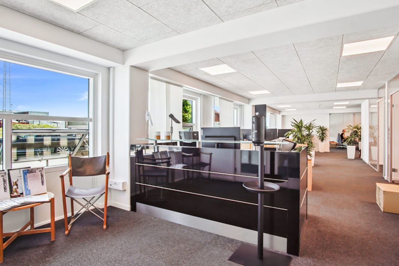 Moderne kontorreception med et sort skrivebord, en stol ved vinduet, indendørs planter og stærkt naturligt lys fra store vinduer.