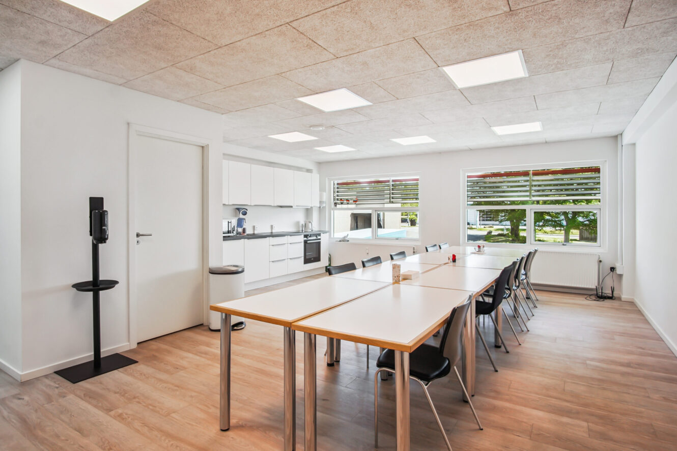 Et moderne pauserum med hvide vægge, et tekøkken, to borde med sorte stole, store vinduer og trægulv.