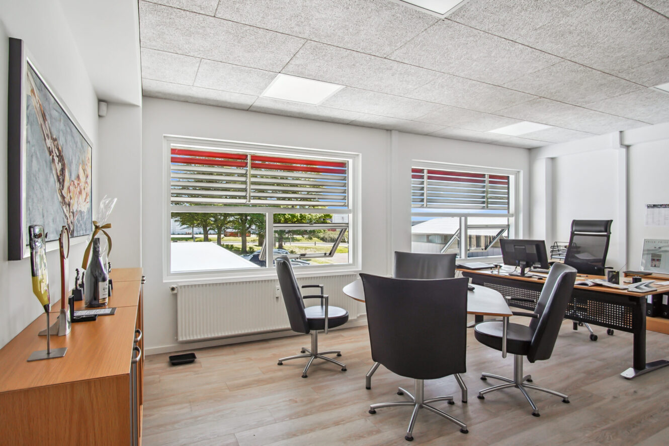 Moderne kontor med store vinduer, et rundt mødebord med stole, et træskrivebord med computere og et sideskab; grønne områder udenfor kan ses gennem vinduerne.