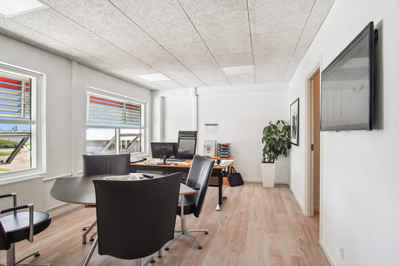 Moderne kontor med trægulv, store vinduer, et skrivebord med computerskærme, et rundt mødebord med stole, et væghængt tv og en potteplante.