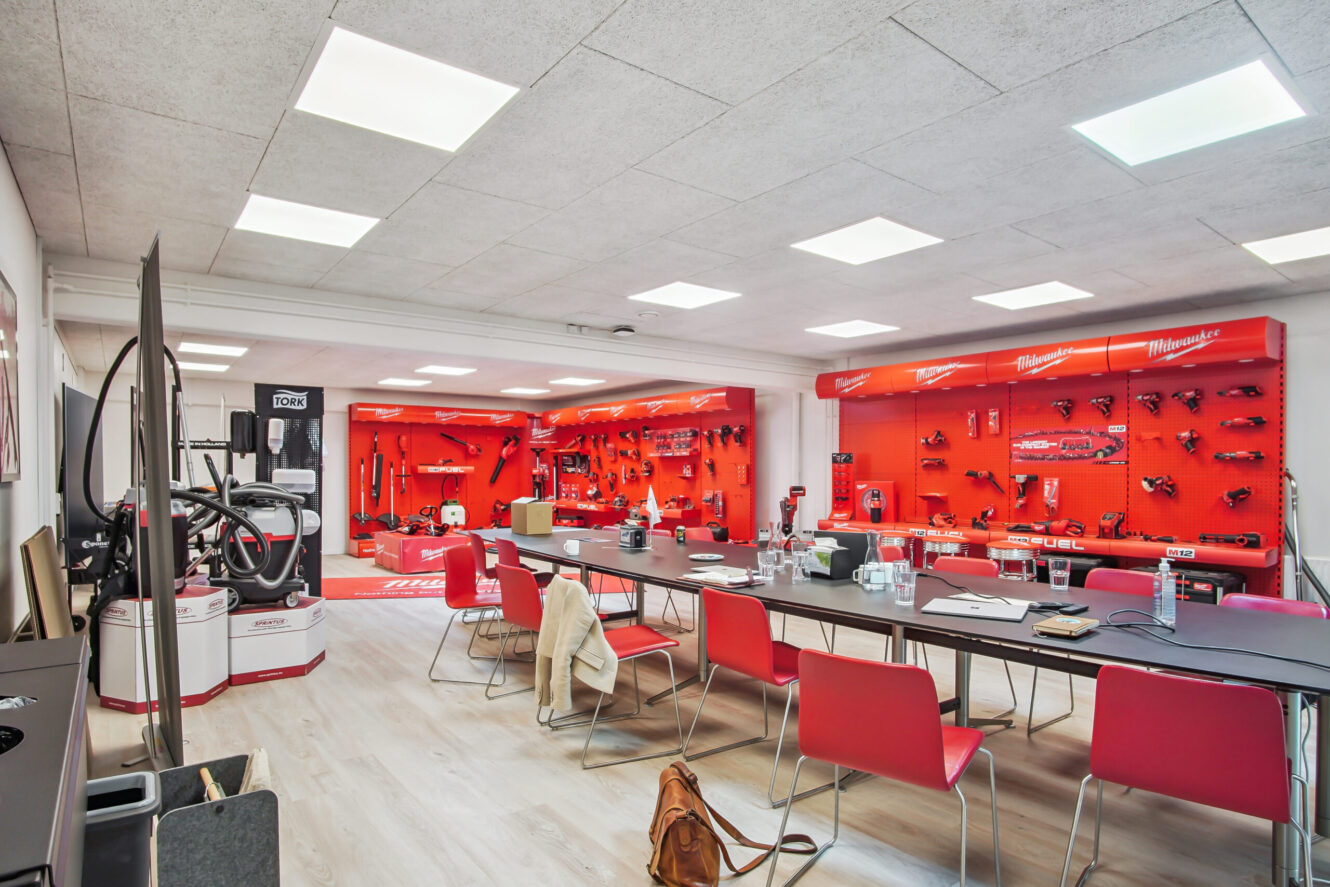 Moderne mødelokale med røde stole, store borde og en vægudstilling med forskelligt elværktøj og udstyr på røde hylder.