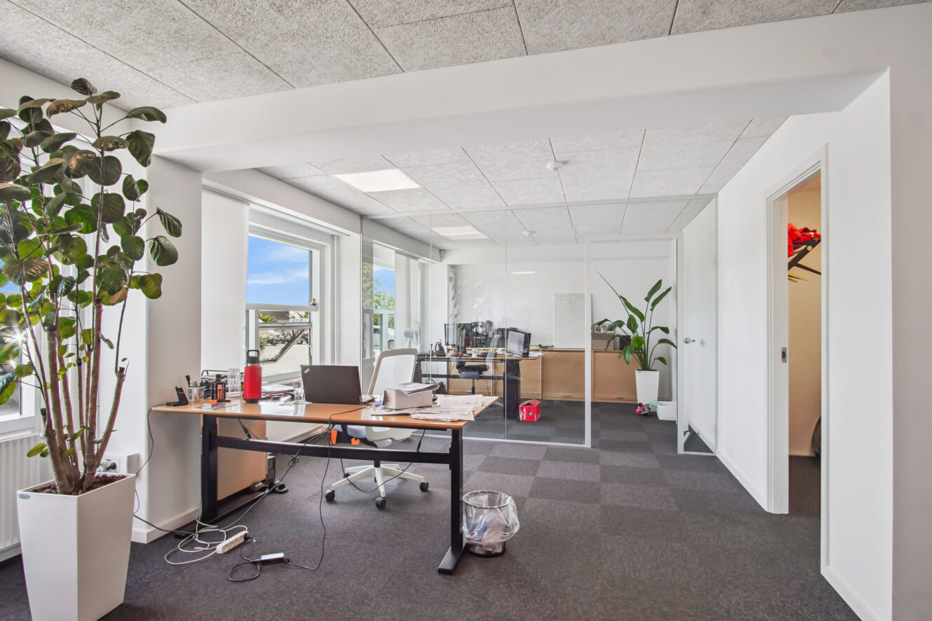 Moderne kontor med skriveborde, stole, potteplanter, store vinduer og glasvægge; papirer og elektronik på et skrivebord med udsigt til et tilstødende kontorområde.