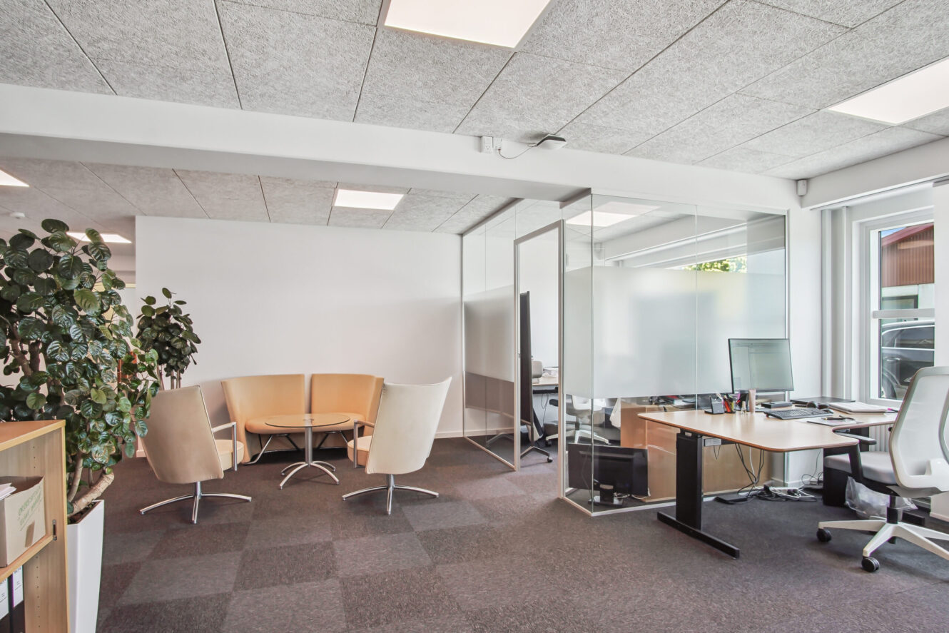 Moderne kontor med et mødeområde med glasvægge, fire beige stole omkring et lille bord, skriveborde med computere, store vinduer og en plante i hjørnet.