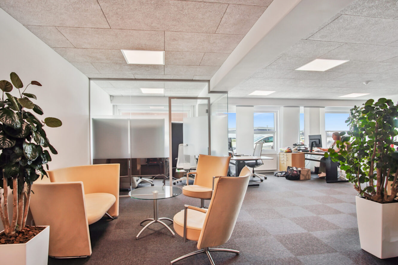 Moderne kontor med beige stole, glasskillevægge, potteplanter og store vinduer; en person arbejder ved et skrivebord i baggrunden.