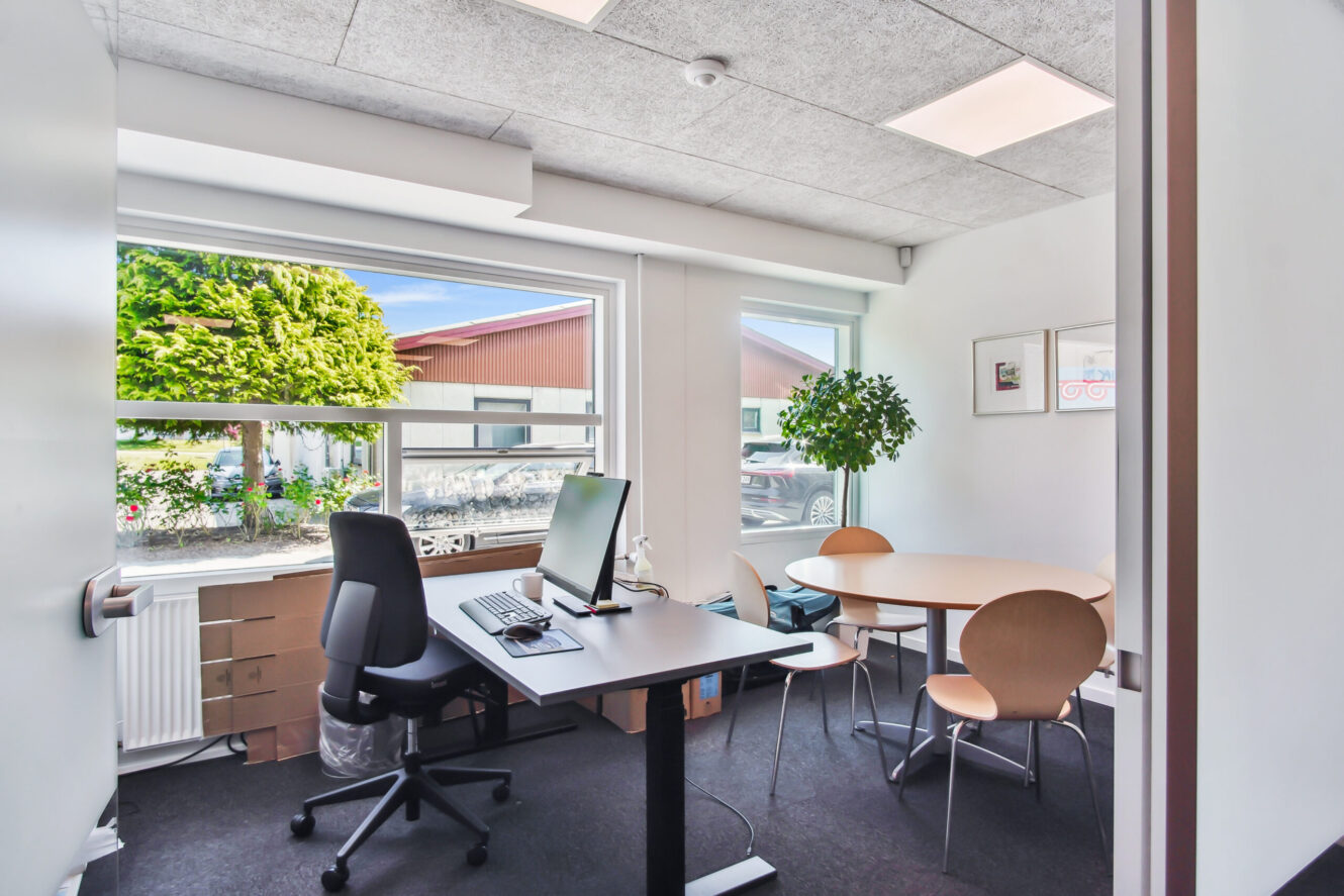 Moderne kontor med skrivebord, computer, stol, rundt mødebord med fire stole, stort vindue, potteplante og udsigt til det fri.