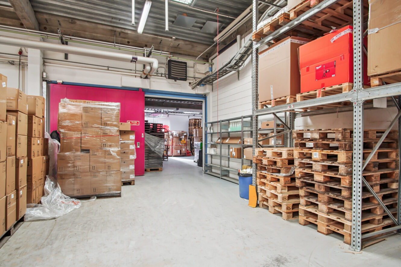 Lageret er indrettet med stablede paller, kasser og metalreoler på et betongulv med en lyserød dør på bagsiden.