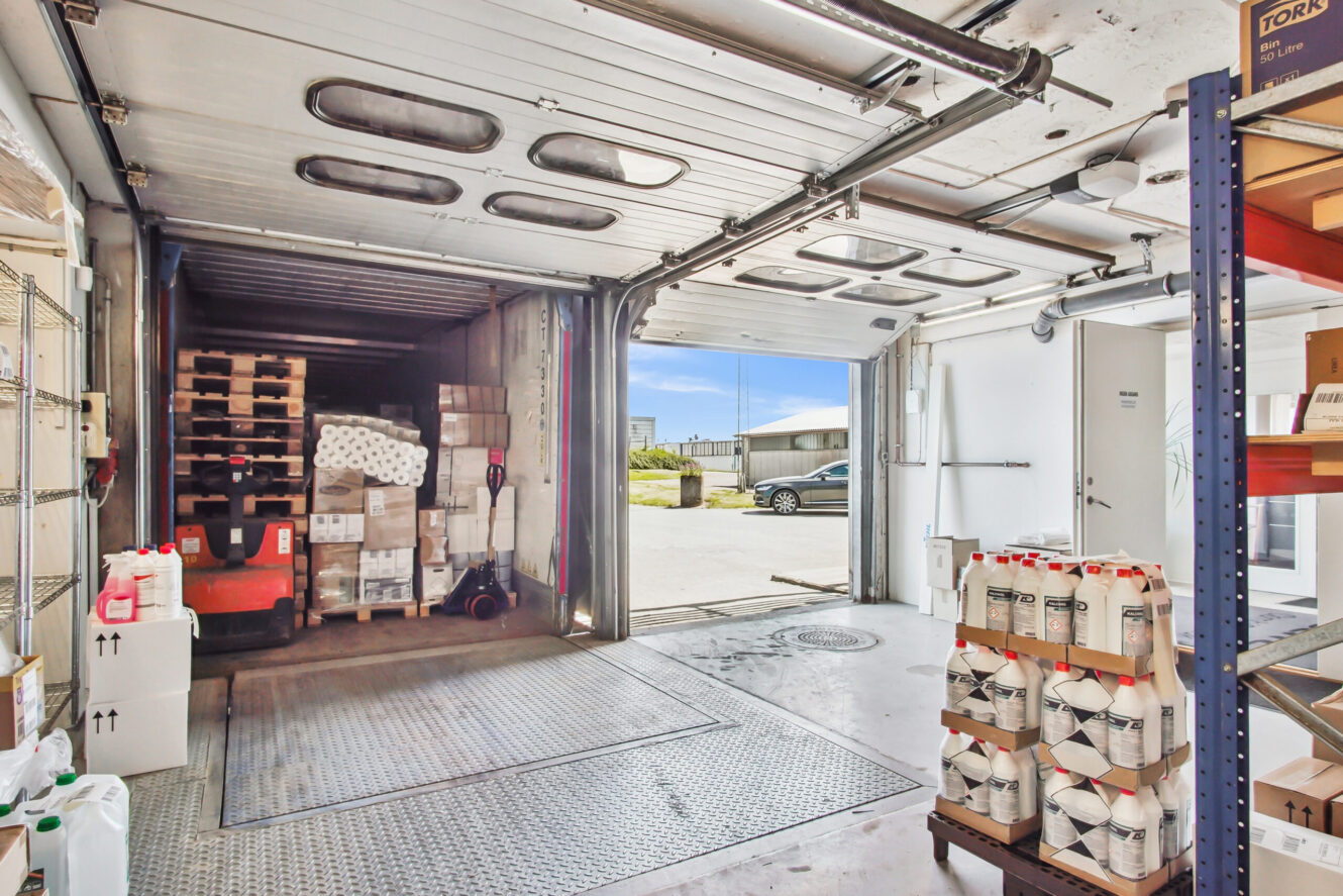 En lagerhal med stablede paller med kasser og rengøringsartikler, en åben garageport og en bil parkeret udenfor under en klar himmel.