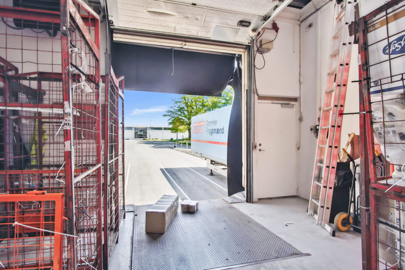 Udsigt fra indersiden af en varevogn med åbne bagdøre, der viser pakker på gulvet, metalvogne, stiger og en mærkevare-trailer parkeret udenfor på en lagerplads.