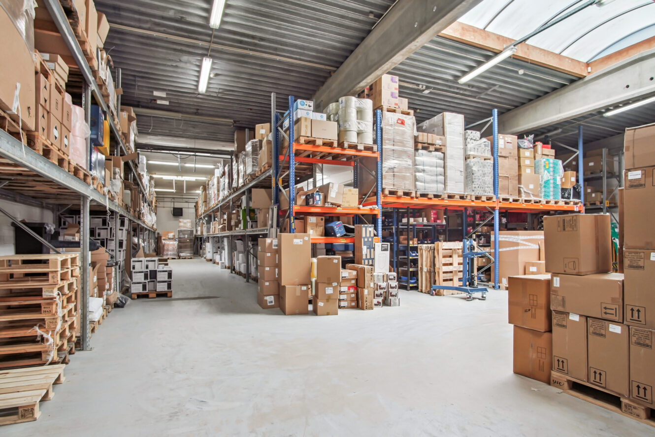 Lageret er indrettet med hylder fyldt med kasser og varer, paller på gulvet og lysstofrør over hovedet.