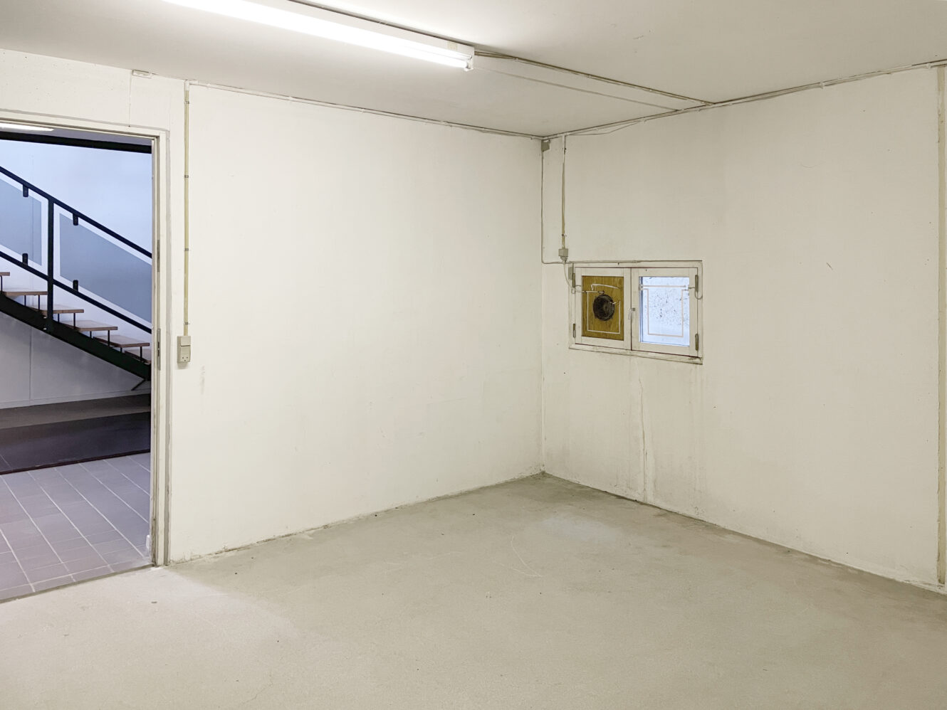 Tomt hvidt rum med et lille firkantet vindue, synlige elektriske ledninger og en åben dør, der fører til en trappe. Lysstofrørsarmatur i loftet.