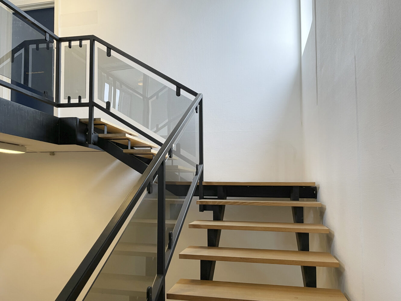 En moderne indendørs trappe med trætrin, sort metalramme og gelænder af glaspaneler, der går op langs hvide vægge og et vindue.