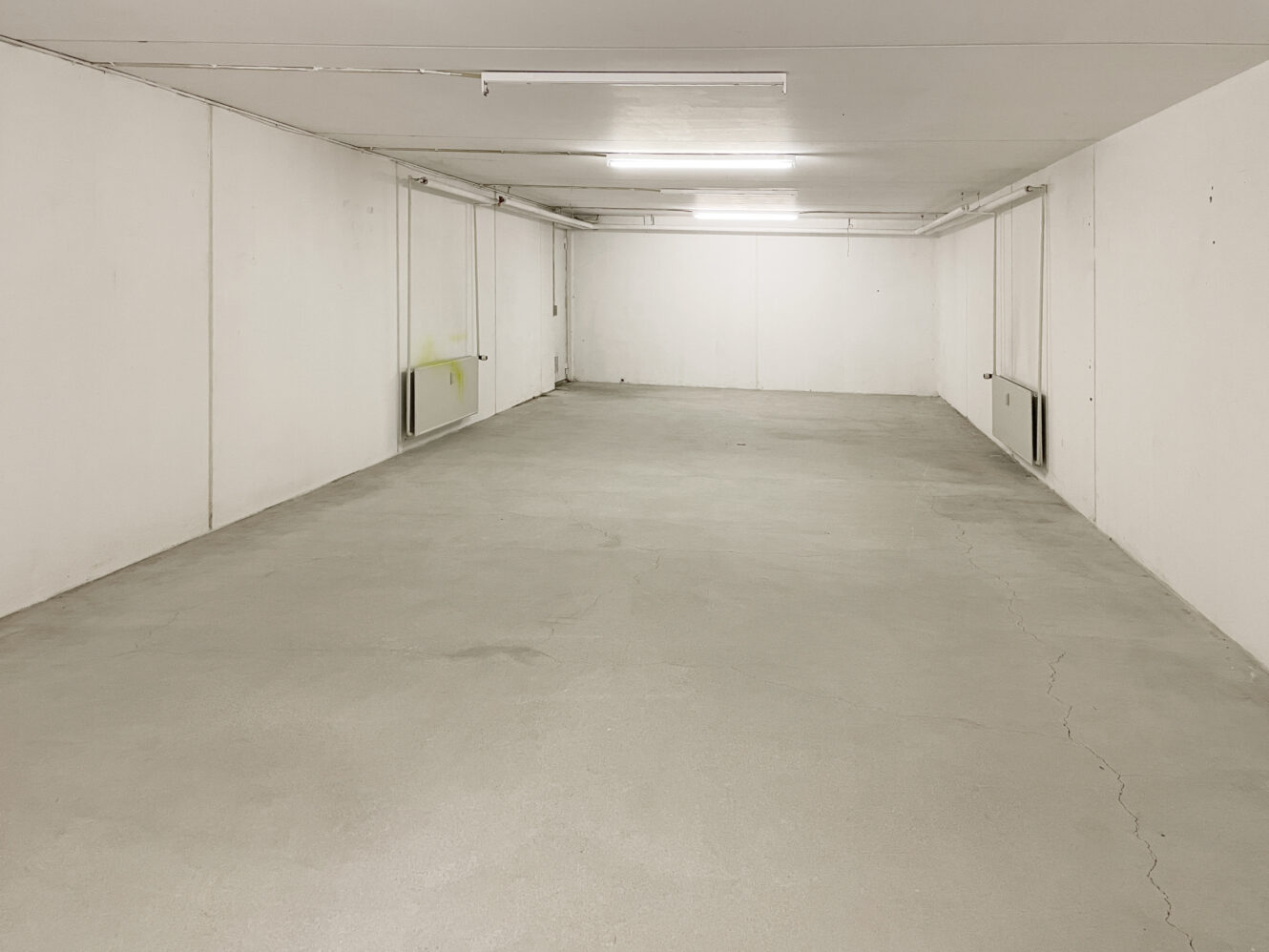 Tom, hvidmuret parkeringskælder eller lagerområde med bart betongulv, lysstofrør i loftet og minimale markeringer eller inventar.