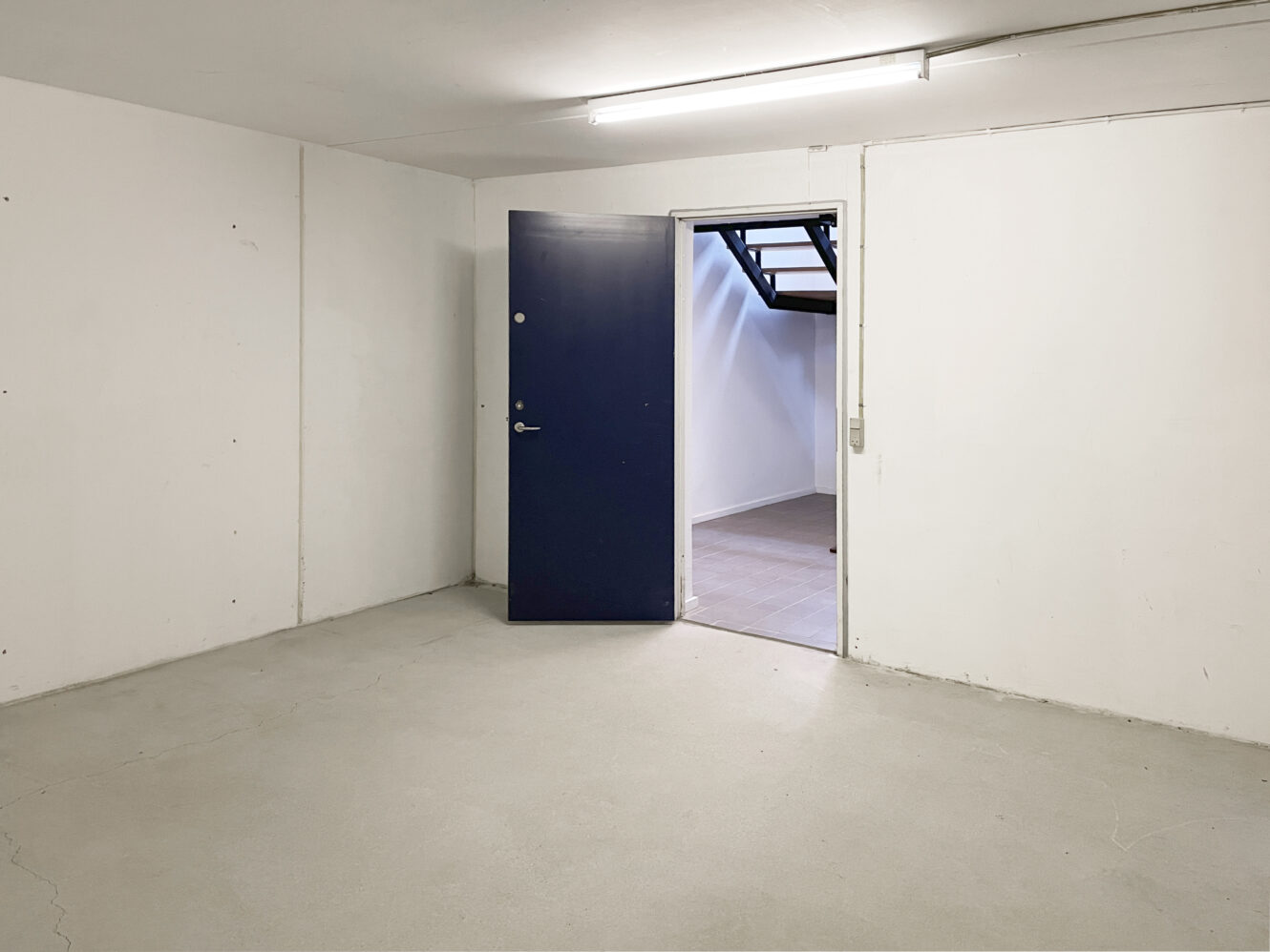 Et tomt rum med hvide vægge og et gråt gulv med en delvist åben blå dør, der fører til et andet oplyst rum med trapper.