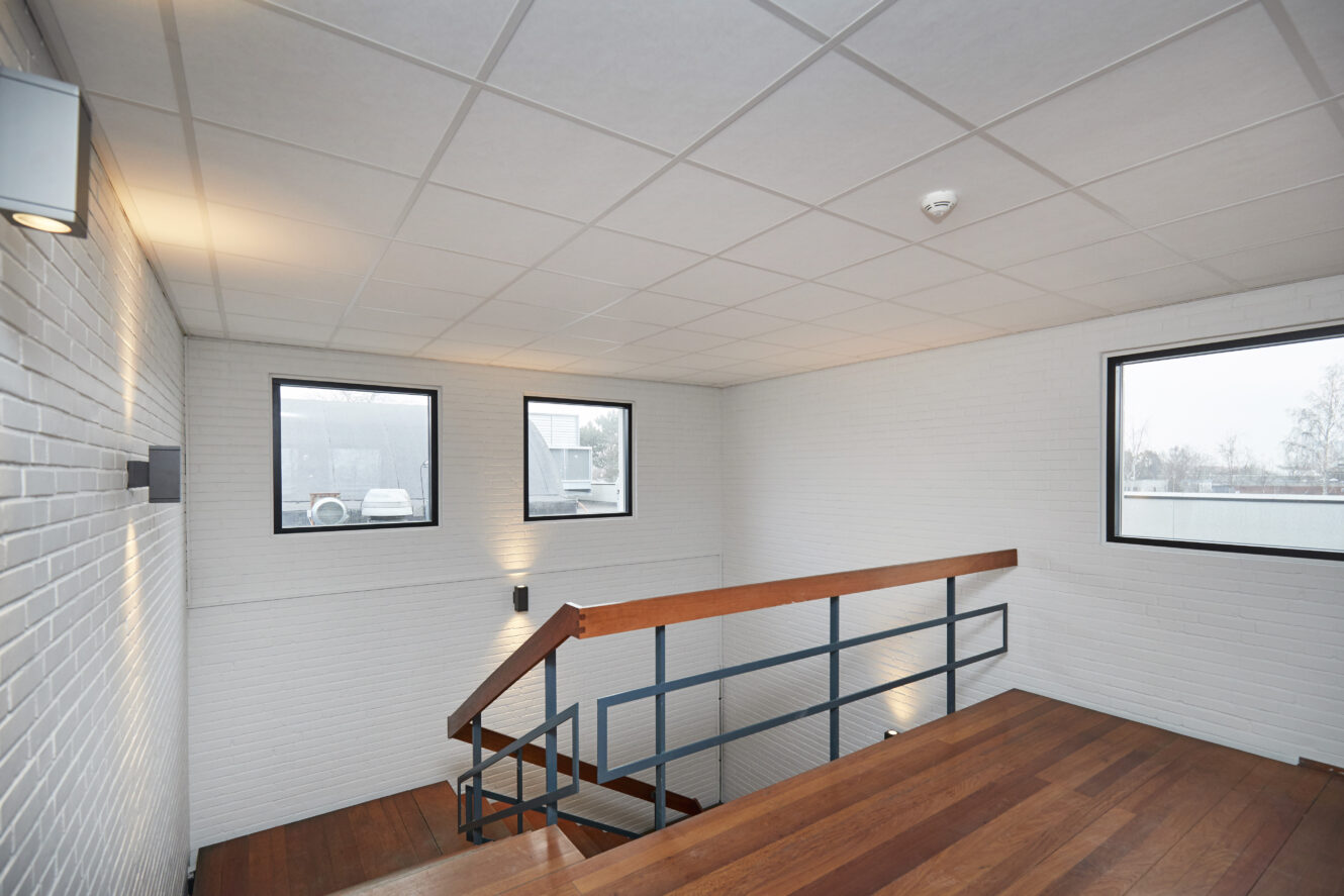 En moderne trappeopgang med trætrin, metalgelænder, hvide murstensvægge og tre firkantede vinduer, der lukker naturligt lys ind.