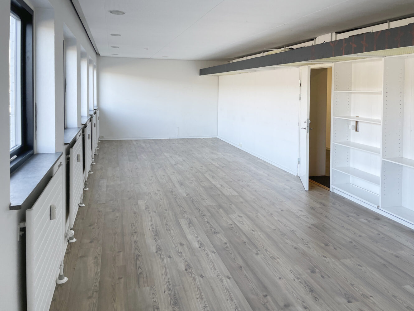 Et langt, tomt rum med lyst trægulv, hvide vægge, flere vinduer til venstre og indbyggede hvide hylder til højre ved en åben dør.