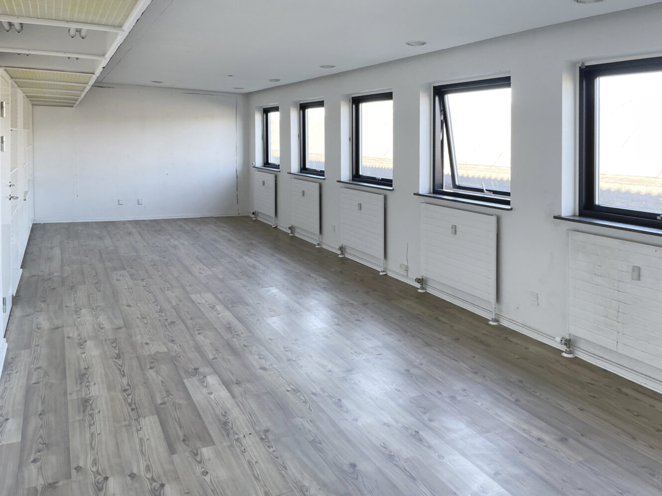 Tomt rum med lyst trægulv, hvide vægge og fem sortindrammede vinduer langs den ene side, der lukker naturligt lys ind.