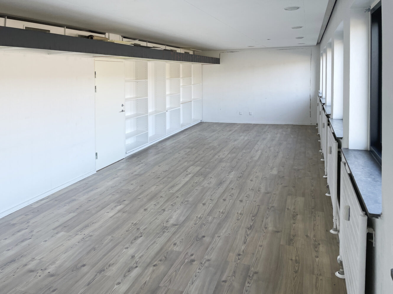 Et stort, tomt rum med lyst trægulv, hvide vægge, indbyggede hylder på den ene side og flere vinduer langs den modsatte væg.