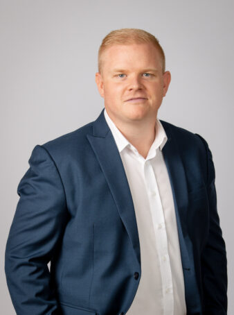 En mand med kort blondt hår i marineblåt jakkesæt og hvid skjorte står på en almindelig lysegrå baggrund og kigger ind i kameraet med et neutralt udtryk.