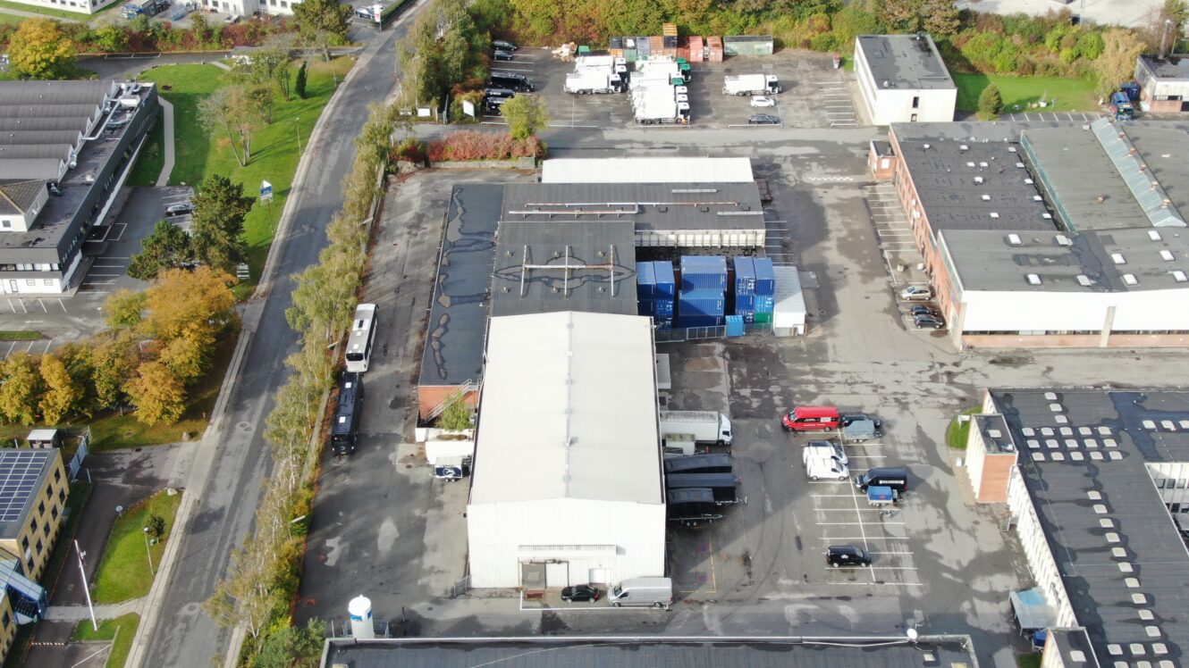 Luftfoto af et industriområde med lagerbygninger, parkerede lastbiler, skibscontainere og flere køretøjer på asfalterede grunde omgivet af nogle træer og bygninger.