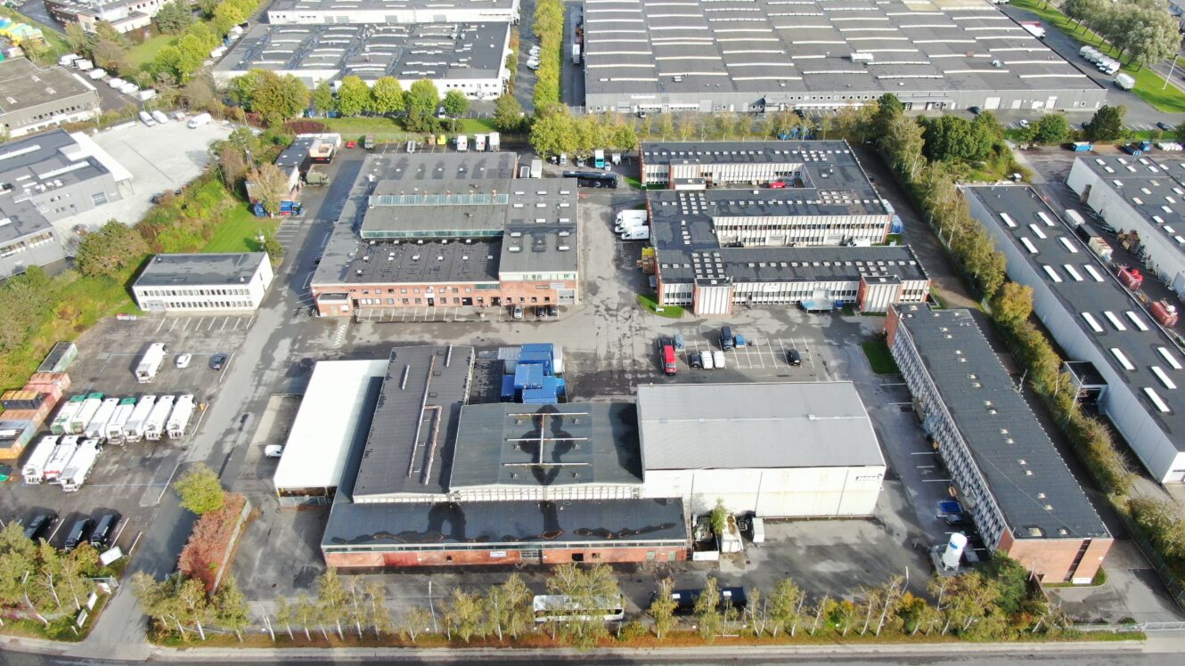 Luftfoto af et industrikompleks med flere lagerbygninger, parkeringspladser og omkringliggende grønne områder.