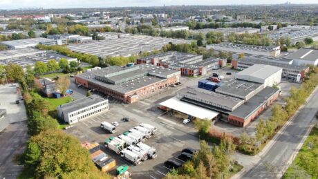 Luftfoto af et industrikompleks med flere lagerbygninger, parkerede lastbiler og omkringliggende grønne områder; et bybillede ses i baggrunden.