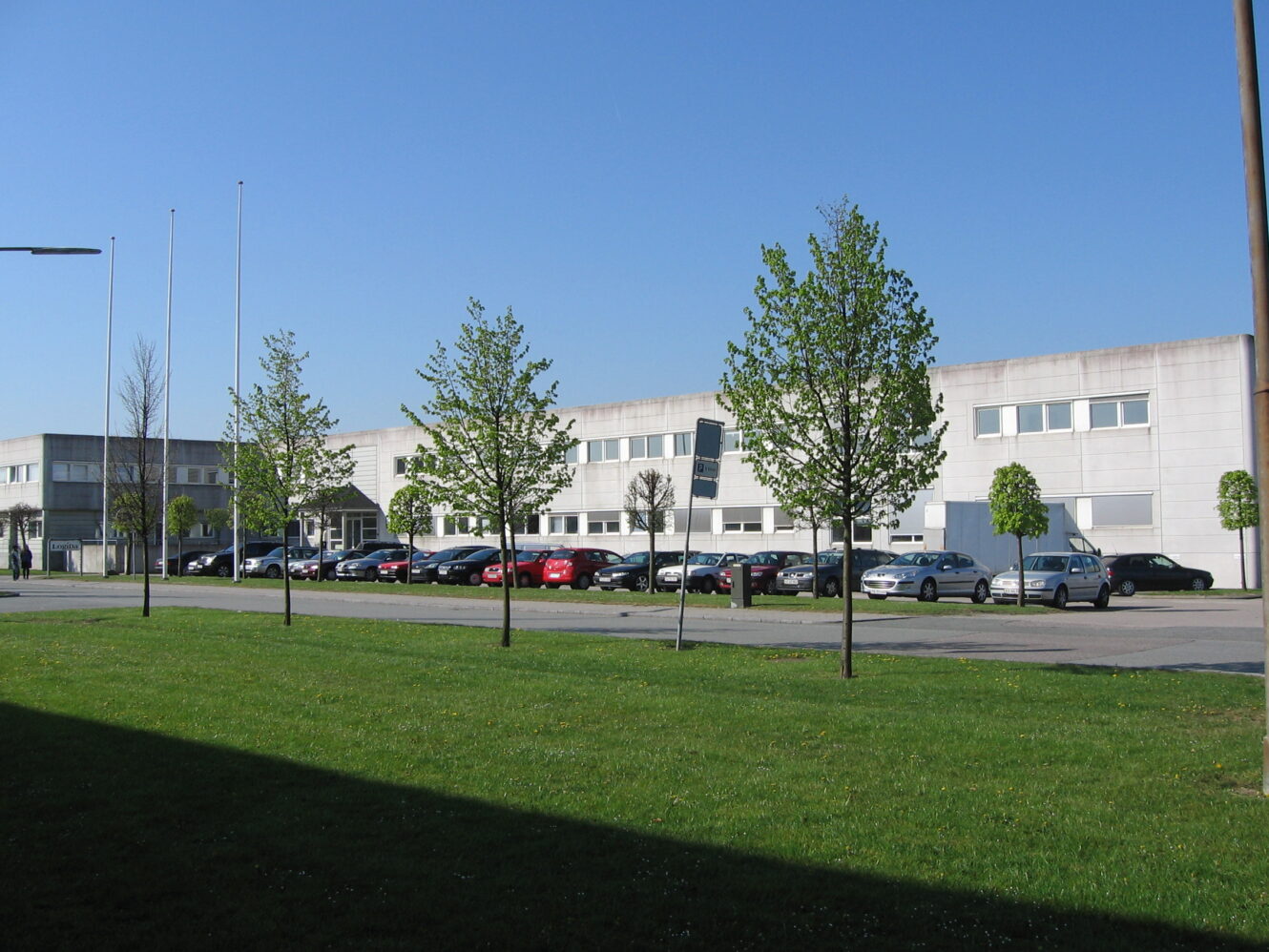 En moderne, toetagers bygning med store vinduer, en række parkerede biler foran og en græsplæne med unge træer i forgrunden under en klar blå himmel.