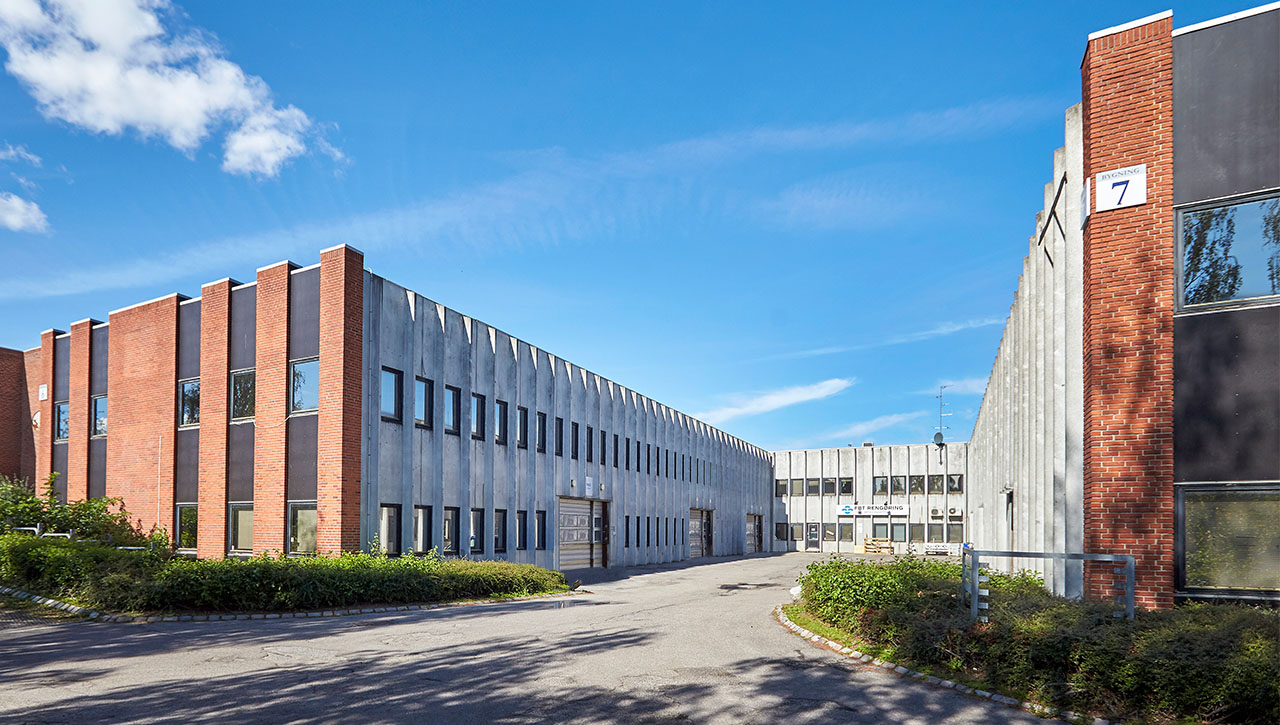 En moderne industribygning med røde mursten og betonvægge, store vinduer og en indkørsel under en klar blå himmel.