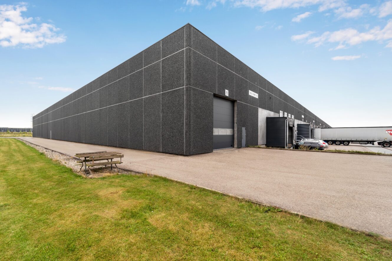 Stort, moderne industrilager med mørkegråt ydre, læsseramper og en asfalteret indkørsel; græsareal og bænk i forgrunden.