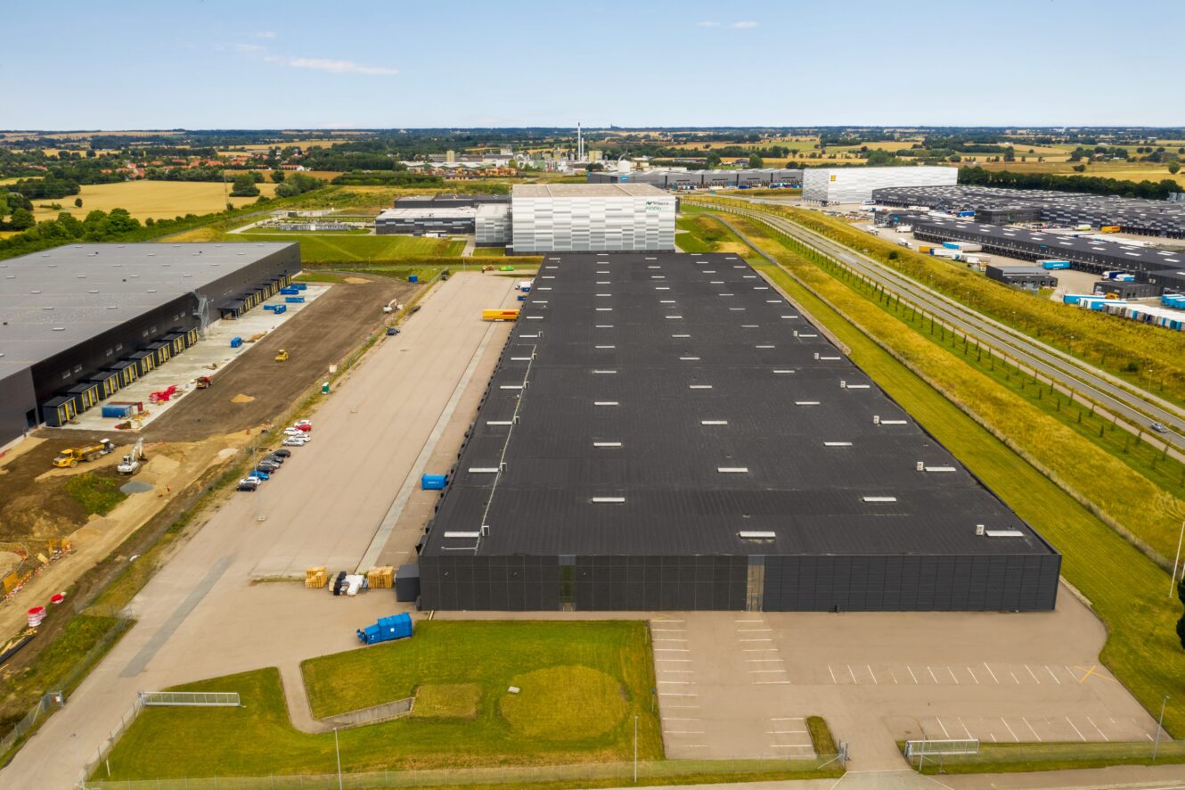 Luftfoto af et stort industrielt lagerkompleks med sorte tage, parkeringspladser og omkringliggende grønne marker.