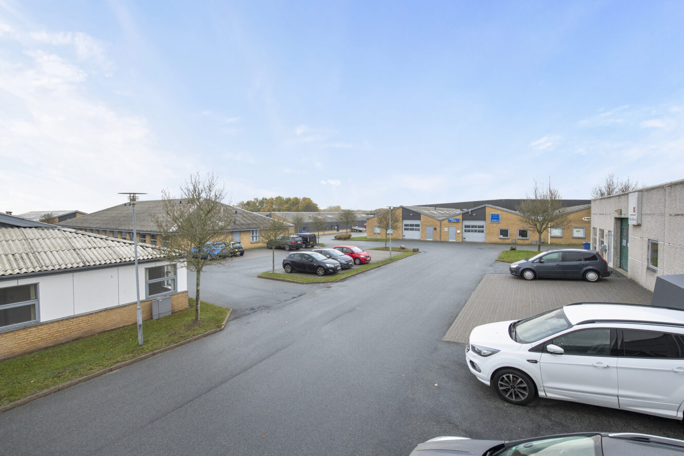 Parkeringsplads med flere parkerede biler omgivet af lave, lyse erhvervsbygninger under en klar himmel.