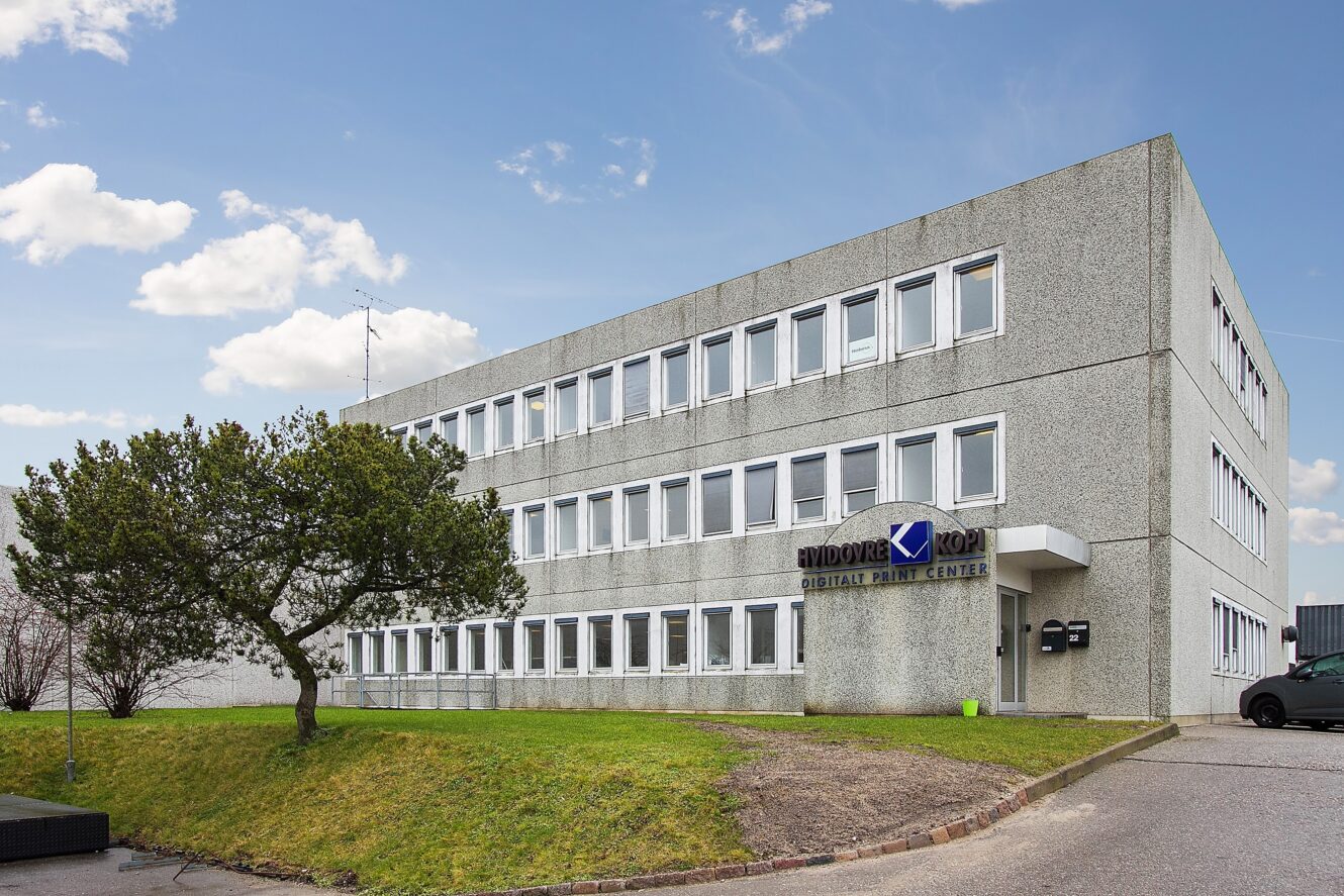 En treetagers grå kontorbygning med mange vinduer, en græsplæne og et træ foran; på et skilt står der Widok Digital Print Center.