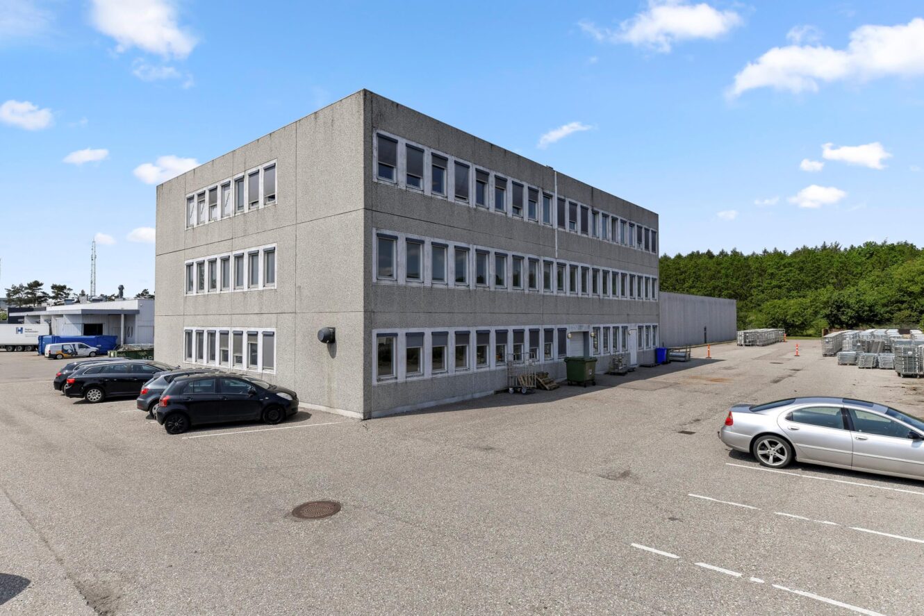 En treetagers grå kontorbygning med mange vinduer, omgivet af parkerede biler og en åben parkeringsplads under en delvis overskyet himmel.