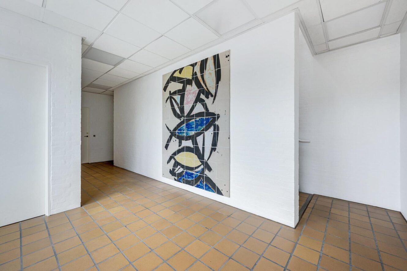 I en hvid gang med lysebrunt flisegulv hænger et stort abstrakt maleri med sorte, gule og blå former på væggen.