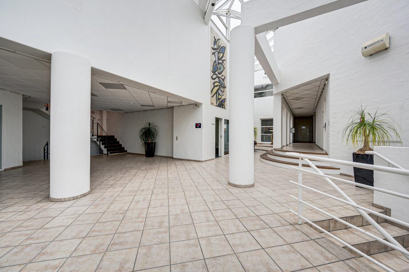 Rummeligt, moderne indendørs atrium med klinkegulve, hvide vægge, søjler, trapper, potteplanter og naturligt lysindfald gennem store vinduer og ovenlys.