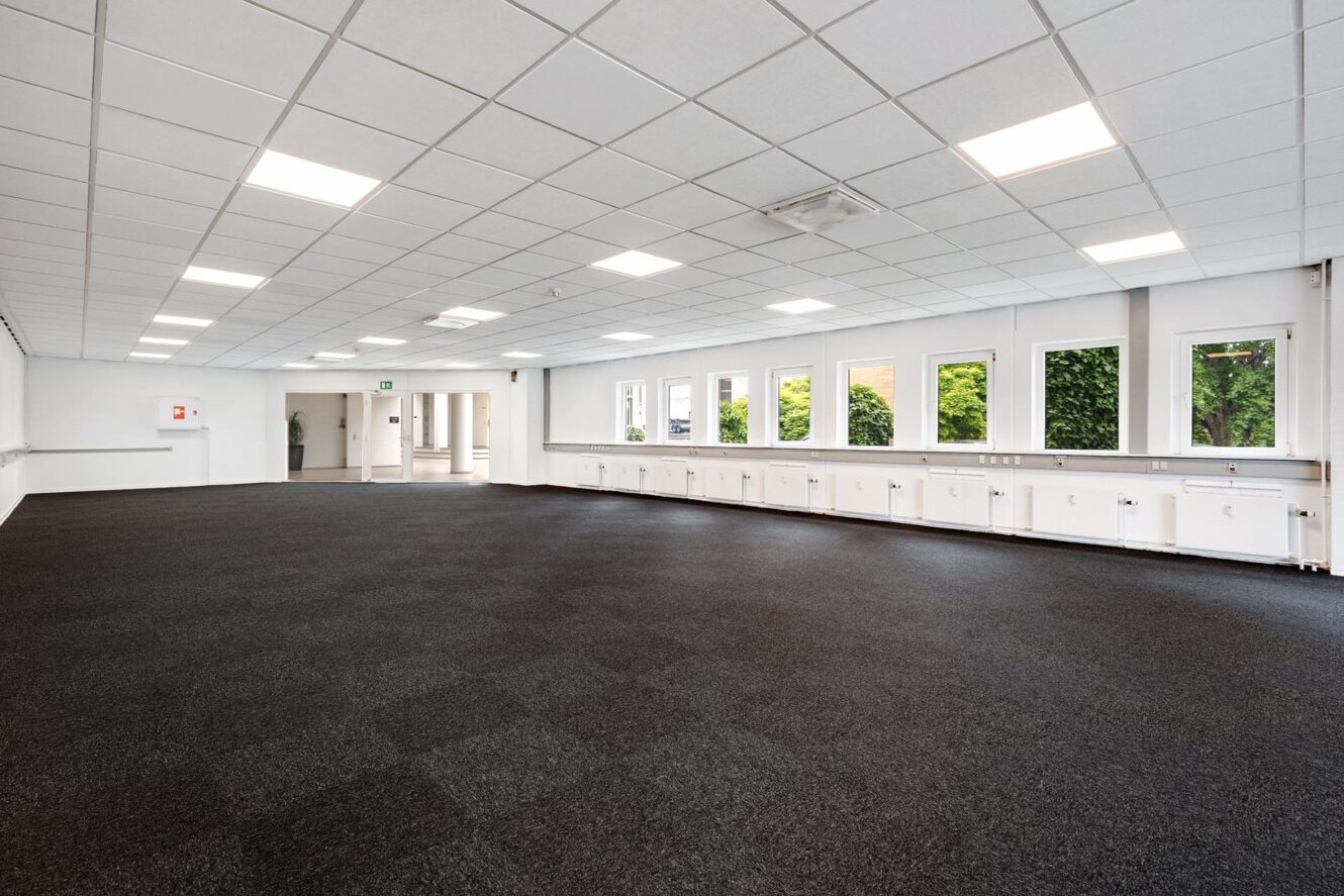 Et stort, tomt rum med sort gulvtæppe, hvide vægge, loftslamper og flere vinduer langs den ene side.