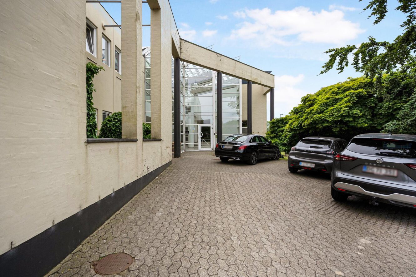 Tre biler holder parkeret i en asfalteret indkørsel ved siden af en moderne, beige bygning med store glasvinduer og en glasindgang.