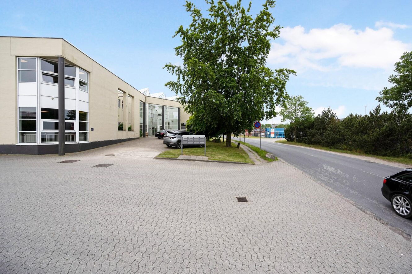 En moderne, beige erhvervsbygning med store vinduer ligger ved siden af en asfalteret vej. Flere parkerede biler og grønne træer er synlige i nærheden under en delvist overskyet himmel.