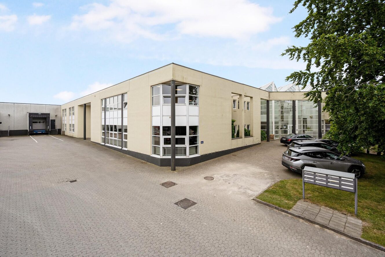 Moderne kontorbygning med store vinduer, en asfalteret parkeringsplads, flere parkerede biler og en grøn græsplæne med træer under en delvist overskyet himmel.