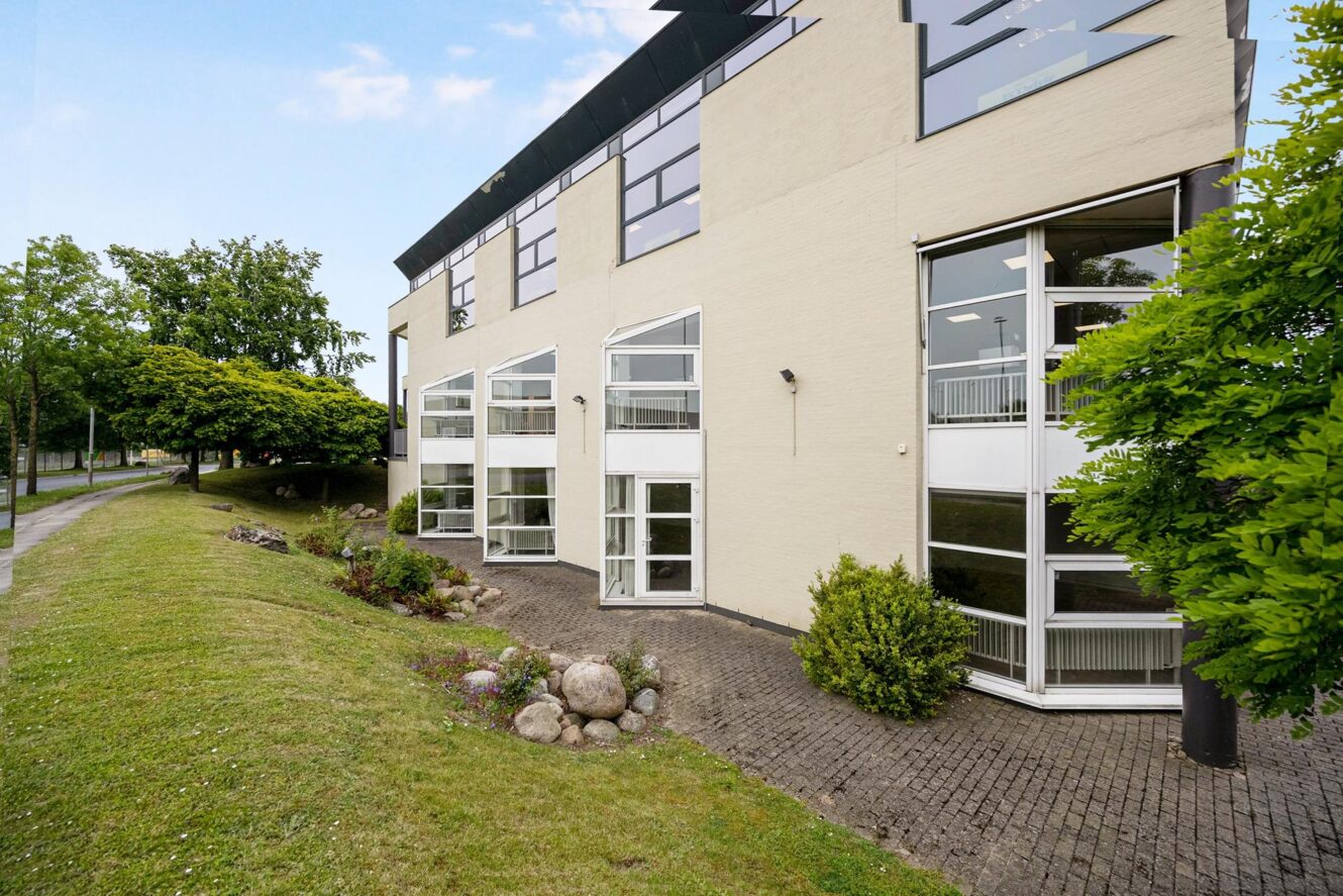 Moderne bygning med store vinduer, lyst ydre og grønne områder omkring en asfalteret gangbro.