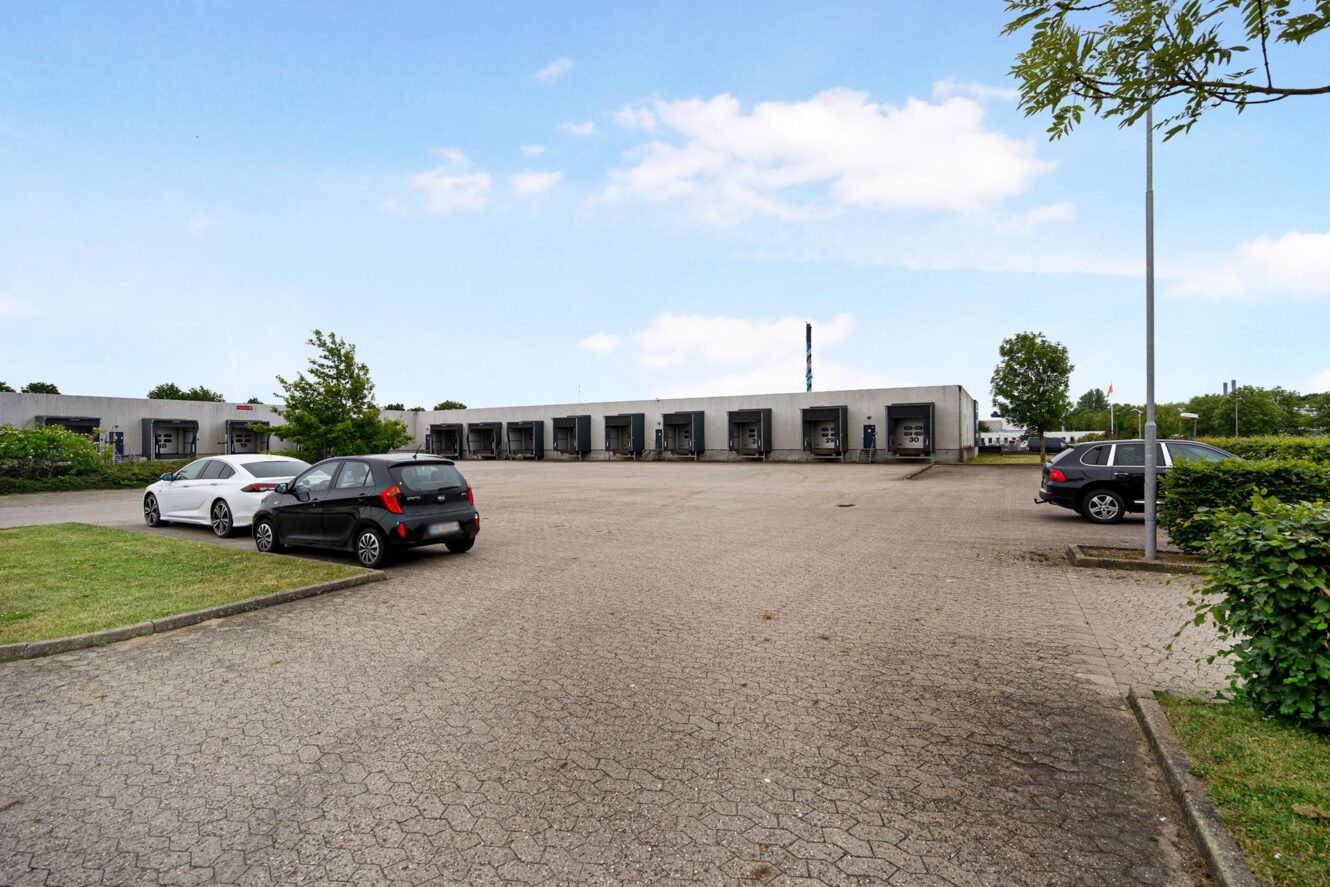 En stort set tom parkeringsplads med nogle få parkerede biler foran en række industrielle læsseramper på en delvis overskyet dag.