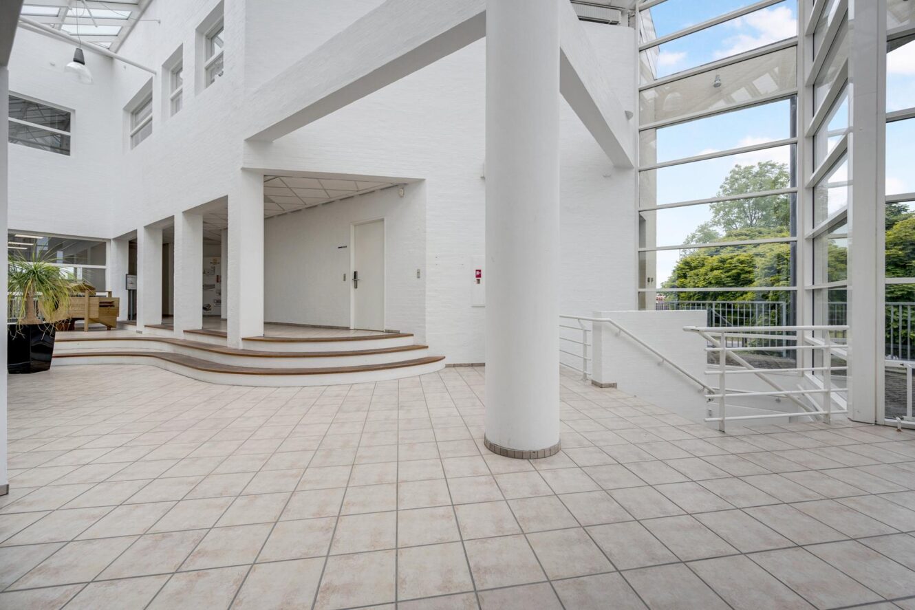 Rummeligt, moderne indendørs atrium med store vinduer, klinkegulv, hvide vægge og flere niveauer, der er forbundet med korte trapper.