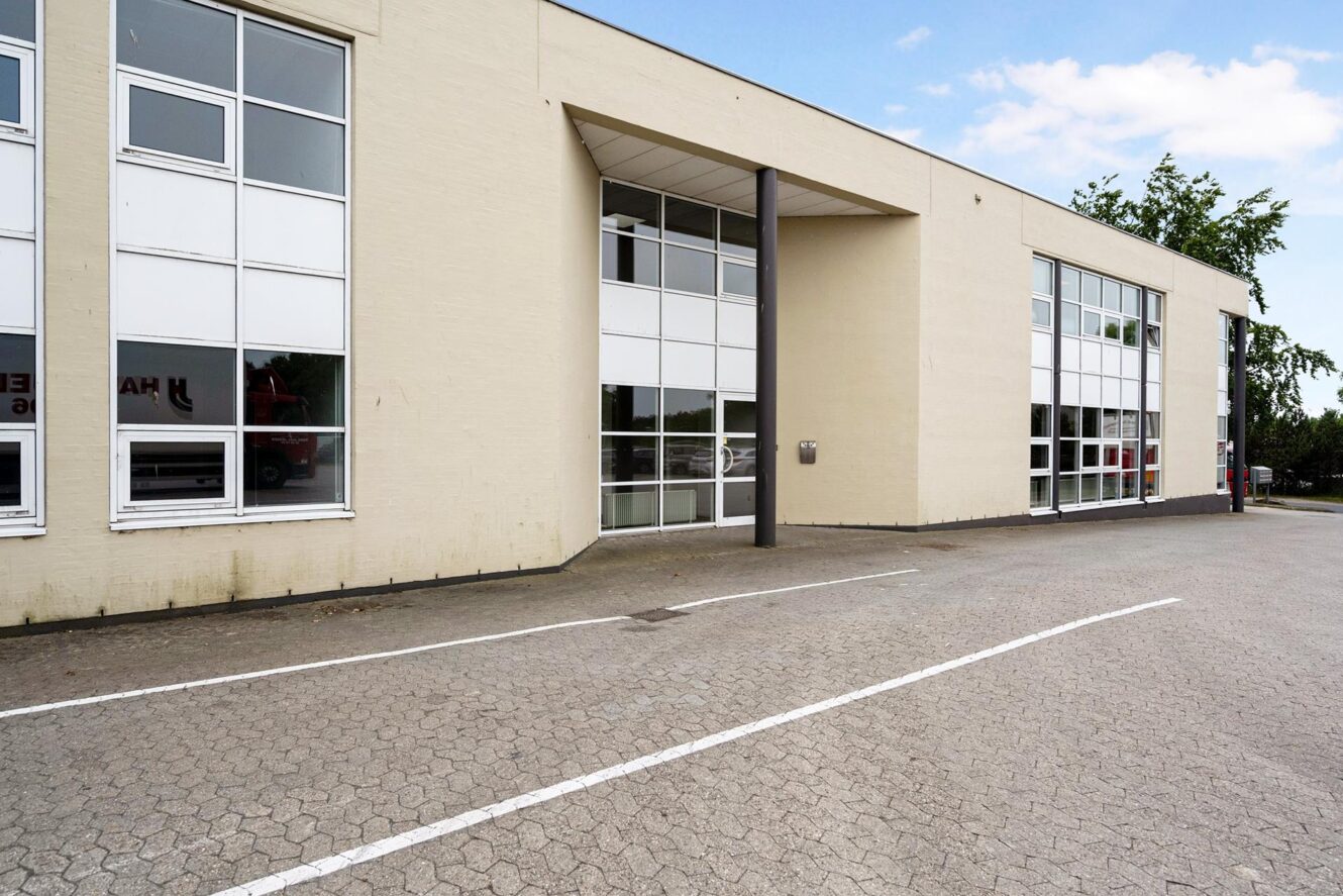 Udvendigt billede af en moderne, beige erhvervsbygning med store vinduer, en indgang under et udhæng og en tom parkeringsplads foran.